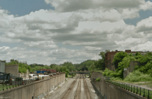 View from Forest Ave. today. Note overgrown bridge abutments where the Tracy Ave. bridge used to be. We assume railroad right-of-way extends approximately from the wall of the building on the left to at least the bridge abutment on the right and possibly to the building out of the picture on the right. This picture provides a better view of the old ramp system bringing tracks up to street level.